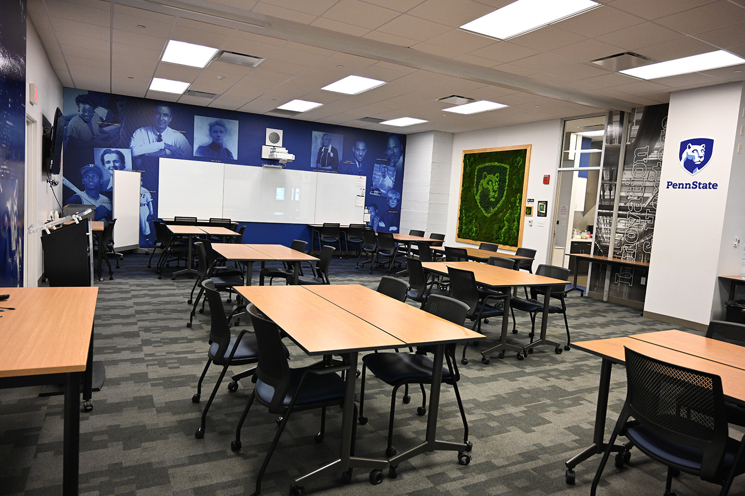 A bright classroom with seven rectangular tables and chairs. There are photos of historical figures on the front wall and a Penn State logo that appears to be made of plants on the right wall.