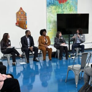 Five professionals sit at the front of a room while speaking on a panel.